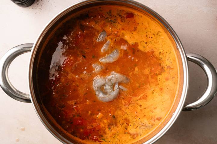 A stainless steel pot on a light surface contains a rich, orange soup with visible chunks of tomatoes and uncooked gray prawns. Steam rises gently, suggesting warmth.