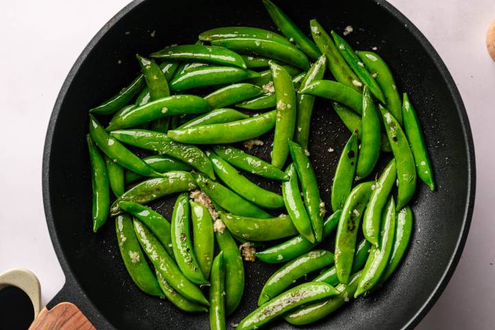 Bright green sugar snap peas are sautéed with minced garlic in a black frying pan, glistening with oil, creating a fresh and savory dish.