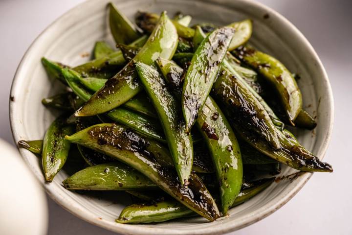 A close-up of glazed sugar snap peas in a white ceramic bowl. The peas are shiny from a savory sauce.