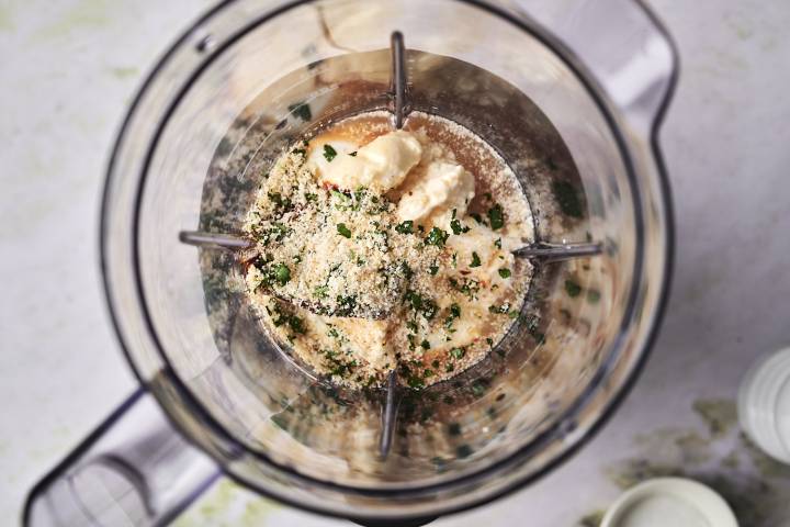 Top view of a blender with ingredients inside, including breadcrumbs, herbs, and a creamy substance.