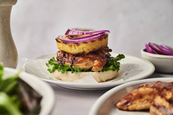 A gourmet chicken sandwich topped with grilled pineapple, red onion, and lettuce on a toasted bun, served with fresh salad.