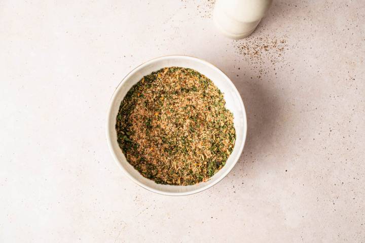 A white bowl filled with a blend of mixed herbs and spices sits on a light speckled surface. A shaker is nearby, suggesting fresh seasoning.