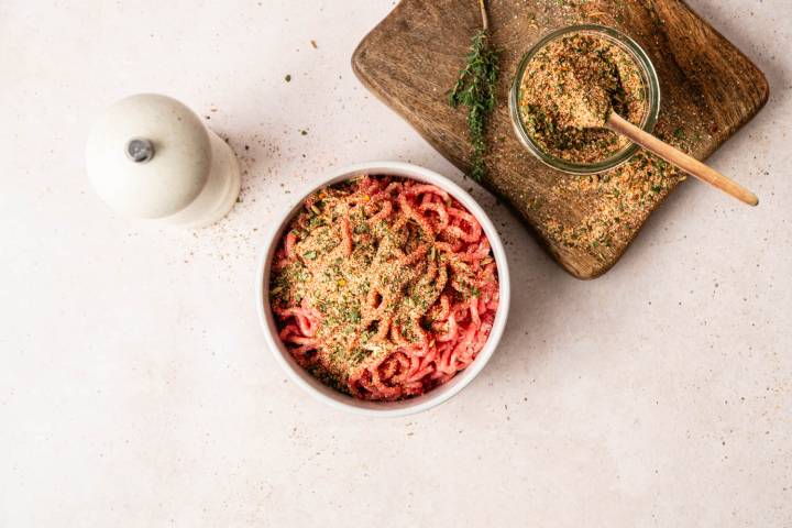 A bowl of raw minced meat topped with seasoning is on a light surface. Nearby is a jar of herbs on a wooden board. A pepper grinder is on the left.
