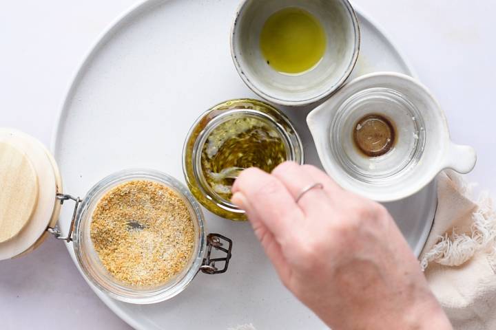 A hand stirs a jar of green pesto on a round white plate with small bowls of olive oil and vinegar, and a jar of breadcrumbs nearby, creating a rustic, culinary scene.