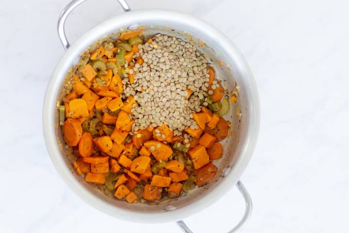 Lentils being added to a soup pot with carrots, celery, and sweet potatoes.