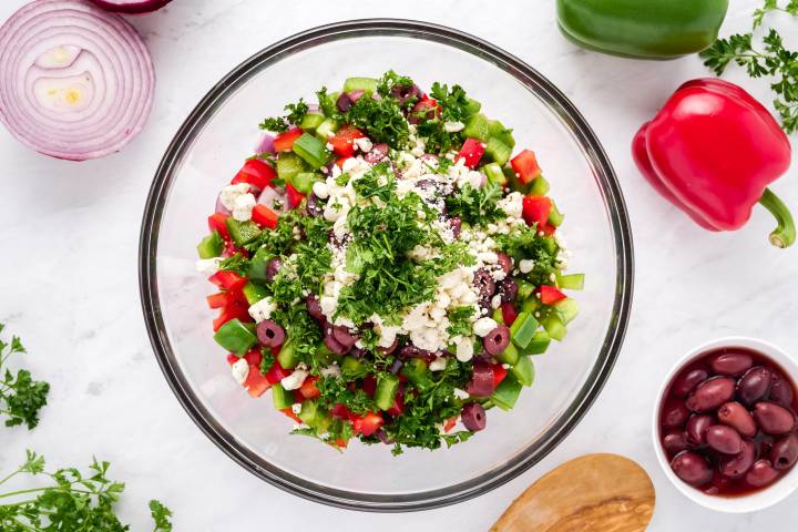 Glass bowl with chickpeas, red onion, cucumber, bell peppers, feta cheese, olives, and parsley.