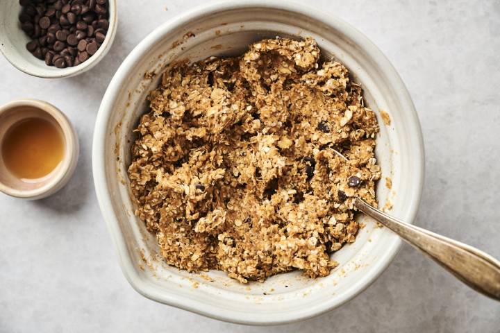 Rolled oats, chia seeds, shreddd coconut, flaxseed meal, and chocolate chips mixed with peanut butter and mashed bananas in a bowl.