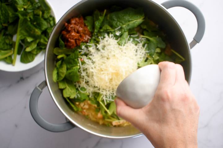 Parmesan cheese being added to a pot with orzo, spinach, white beans, sundried tomatoes, and chicken.
