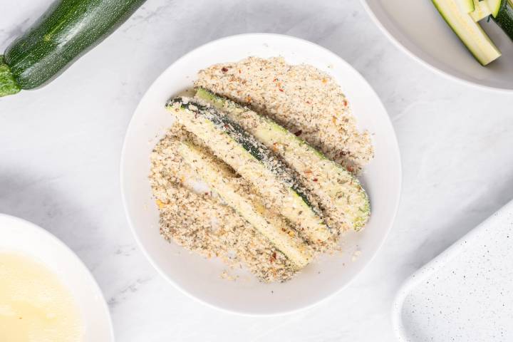 Zucchini slices being dredged in parmesan cheese and breadcrumbs.