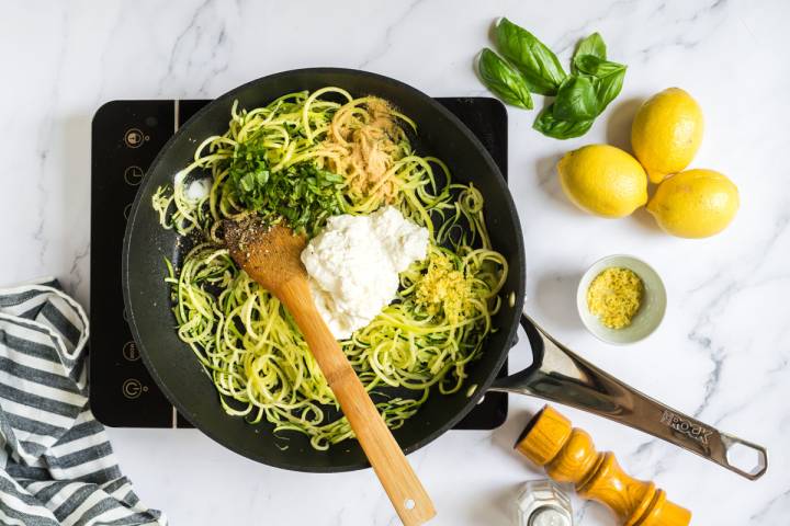 Ricotta cheese, lemon zest, and basil being added to zucchini noodles in a skillet.