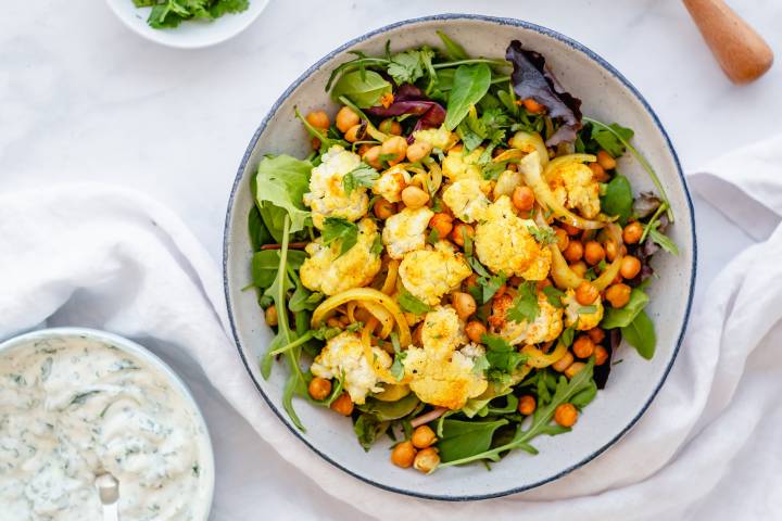 Roasted chickpea and cauliflower in a bowl with greens with yogurt sauce on the side.