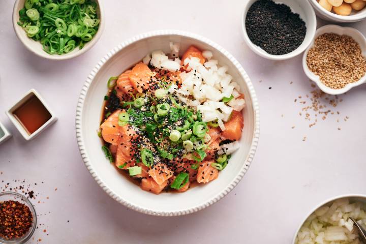Raw salmon cubes with onion, green onions, sesame seeds, soy sauce, rice vinegar, and sesame oil in a bowl.