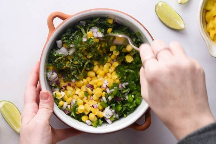 Corn, mango, red onion, cilantro, and lime juice being stirred in a white bowl.