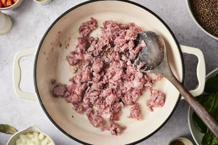 Raw ground sausage being cooked and crumbled in a white Dutch oven with a wooden spoon
