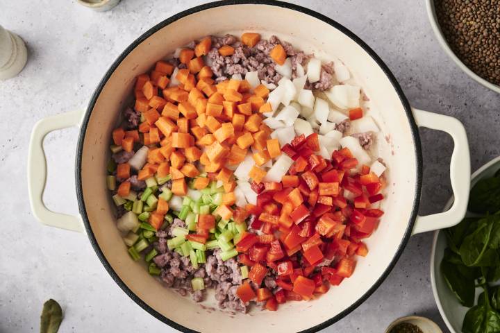 Chopped carrots, celery, onions, and red bell peppers layered over browned sausage in a white Dutch oven, ready to be sautéed for lentil soup.