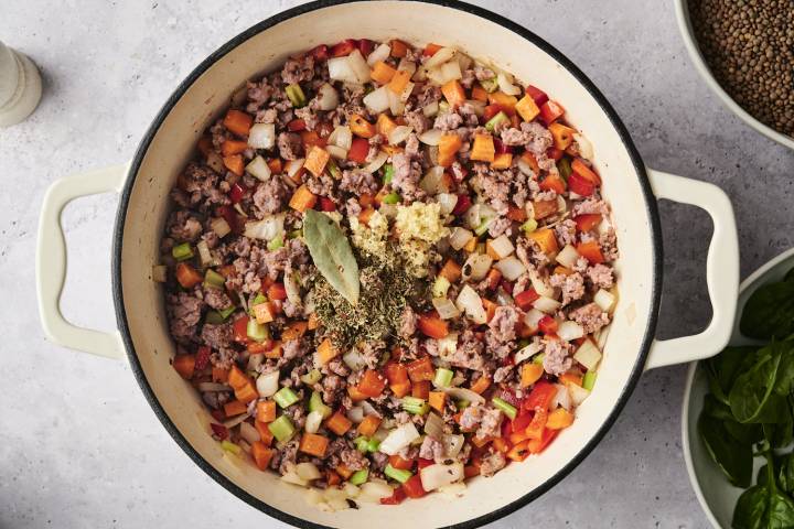 A white Dutch oven filled with browned sausage, diced carrots, celery, onions, and red bell peppers, topped with minced garlic, dried herbs, and a bay leaf, ready to be stirred together for sausage lentil soup.