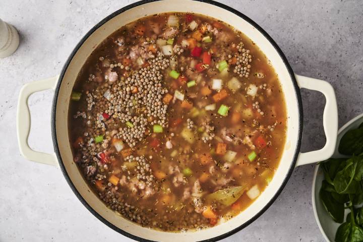 A large pot filled with sausage lentil soup in progress, showing diced carrots, celery, onions, red peppers, and lentils simmering in broth with herbs and bay leaves.