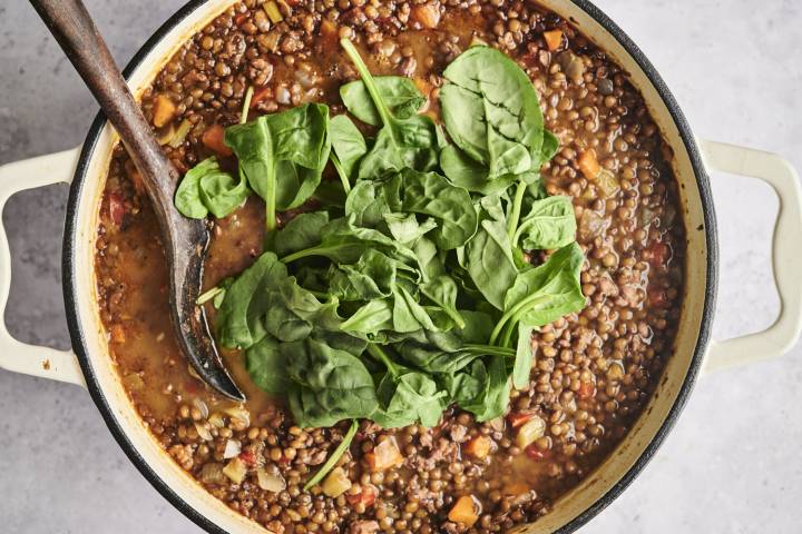 A pot of sausage lentil soup simmering on the stove, topped with fresh spinach leaves before being stirred in, with a wooden spoon resting in the pot.
