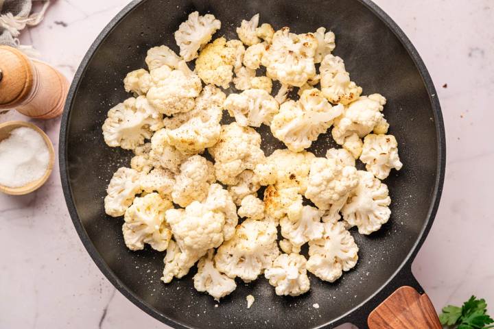 Overhead view of roasted cauliflower florets in a black skillet, with a light sprinkling of pepper. Nearby are a wooden pepper grinder and a bowl of salt on a marble surface.