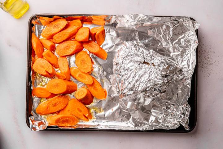 Sliced carrots on foil-lined baking sheet, ready for roasting. Carrots on the left; foil-wrapped section on the right. Oil bottle at the top left.