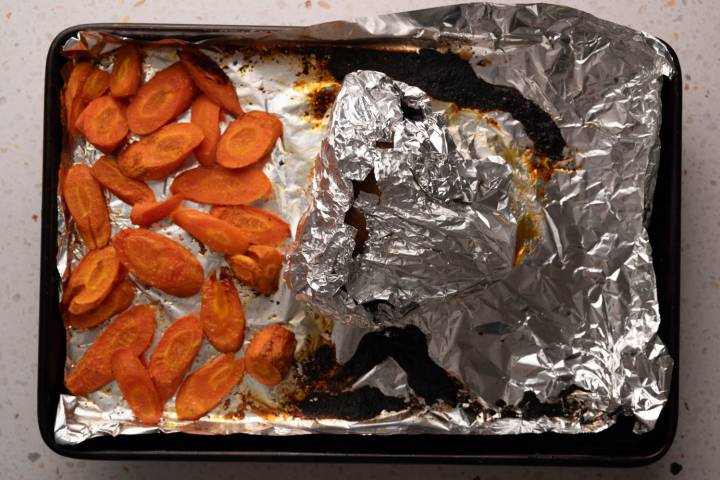A foil-lined baking tray holds roasted orange carrot slices on the left, with a crumpled foil packet on the right, set against a light countertop.