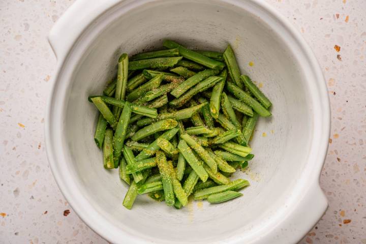 Chopped green beans coated with seasoning in a white ceramic bowl on a speckled countertop.