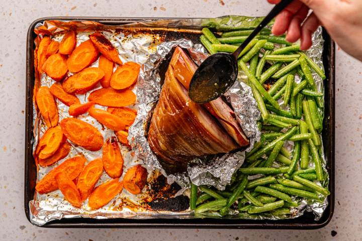 A baking tray lined with foil holds roasted carrots, a ham wrapped in foil, and green beans. A hand drizzles glaze over the ham with a spoon.