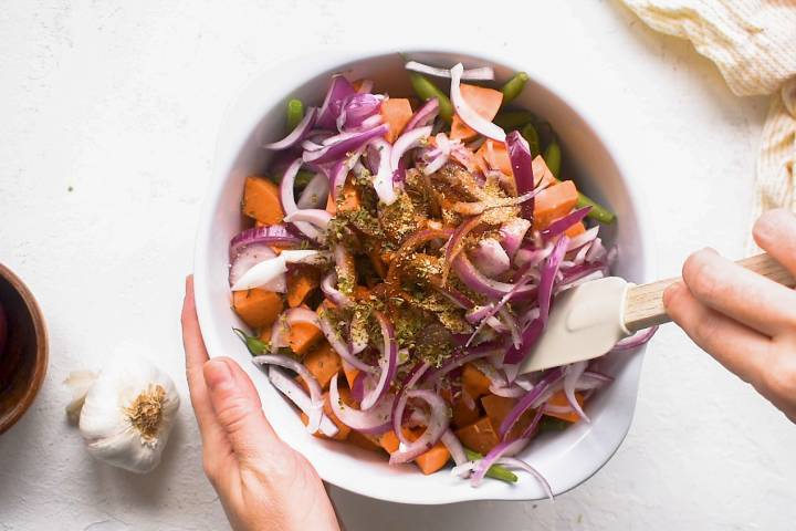 Green beans, sweet ptoatoes, and red onions in a mixing bowl with olive oil and spices.