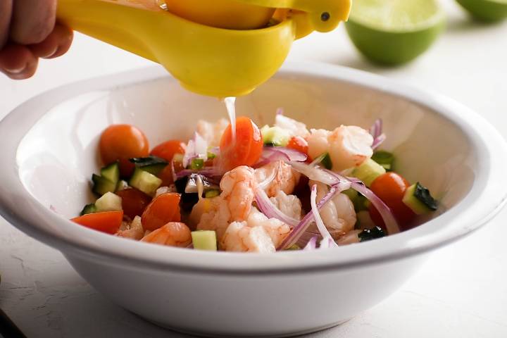 Lime juice being added to chopped shrimp, red onion, cucumber, and tomatoes in a bowl.