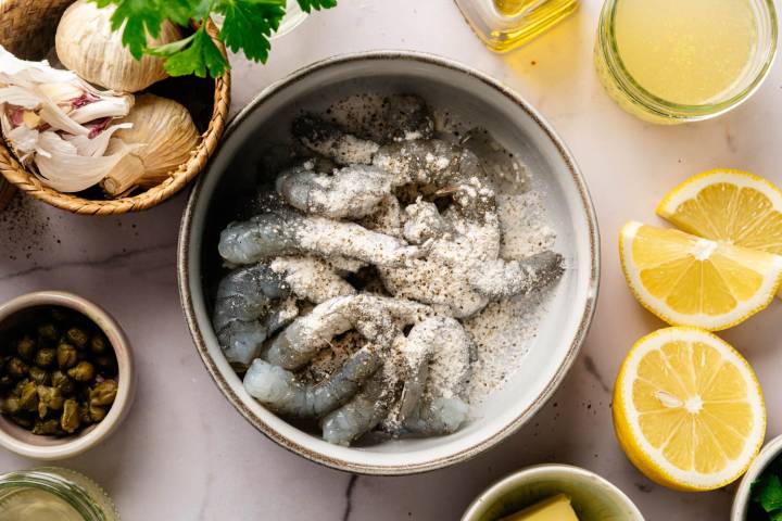 A bowl of raw shrimp seasoned with flour and pepper is surrounded by garlic, parsley, lemon slices, capers, olive oil, and broth on a marble countertop.