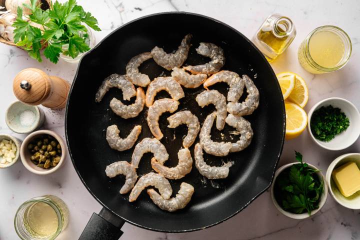 Raw shrimp are arranged in a black pan on a marble counter. Surrounding ingredients include lemon, butter, parsley, garlic, capers, salt, pepper, oil, and broth.