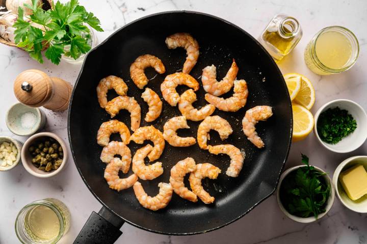 Shrimp sautéing in a skillet, surrounded by ingredients like garlic, capers, lemon, parsley, butter, olive oil, and seasonings on a marble counter.