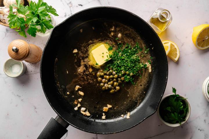 A frying pan with melted butter, capers, chopped garlic, and parsley on a marble countertop. Nearby are lemon wedges, olive oil, and salt. Fresh and vibrant.