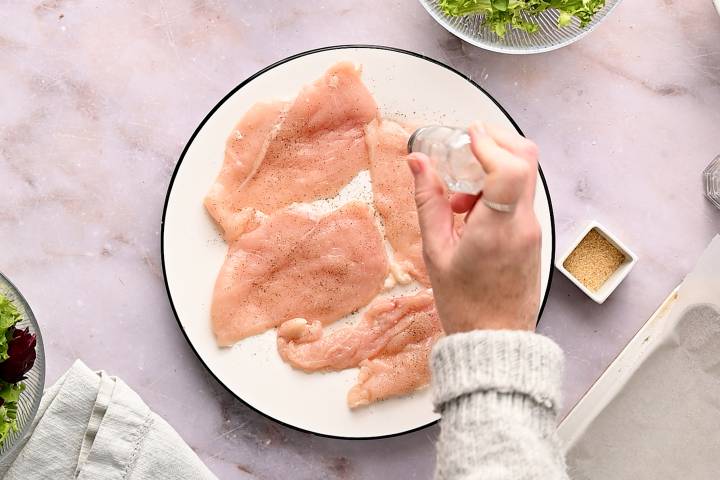 Salt and pepper being sprinkled on chicken breast cutlets.
