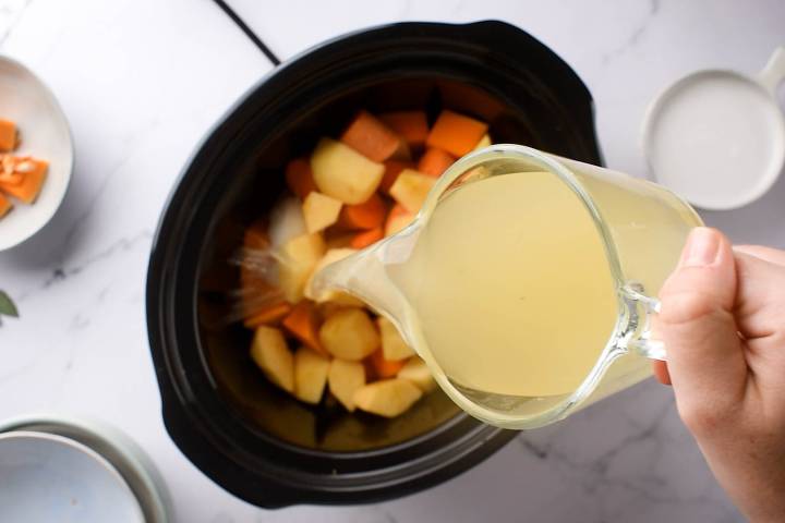 Vegetable borth being poured over butternut squash, apples, carrots, and onions in a slow cooker.