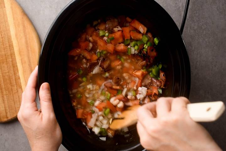 Pinto beans with vegetables, broth, and spices being stirred in a slow cooker.