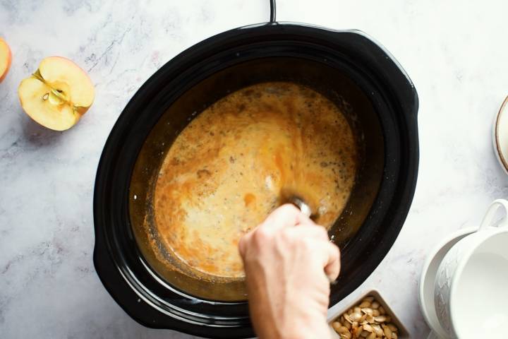 Steel cut oats, almond milk, canned pumpkin, spices, chia seeds, and flaxseeds being stirred in a slow cooker.