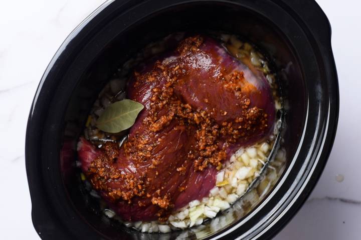Tri-tip covered with spices in a slow cooker with beef broth and a bay leaf.