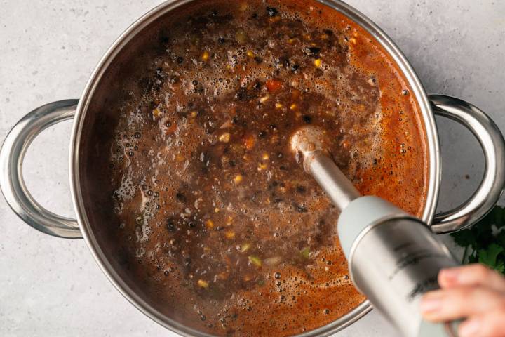 A hand holds an immersion blender blending a hearty black bean soup in a large pot. The soup has visible beans and vegetables, creating a rich texture.