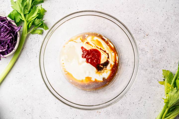 A glass bowl with creamy dressing and red sauce, surrounded by celery stalks and shredded purple cabbage on a speckled white surface. Fresh and vibrant.