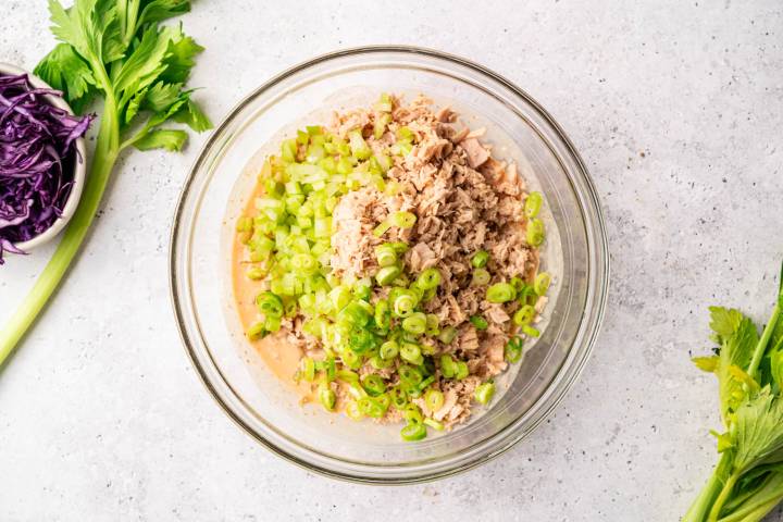 A clear bowl with a fresh tuna salad mixture, featuring diced celery and chopped green onions, sits on a light surface. Nearby are celery stalks and shredded purple cabbage, adding a fresh, vibrant touch.