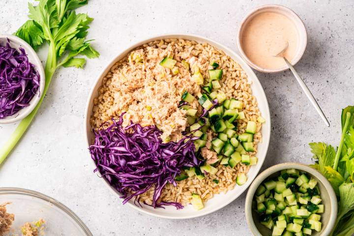 A bowl filled with brown rice, tuna, purple cabbage, and diced cucumbers. Nearby are fresh celery stalks and bowls of sauce and extra cucumbers.