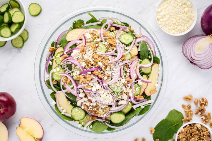 Baby spinach, apples, red onions, cucumbers, walnuts, and goat cheese in a large glass bowl.