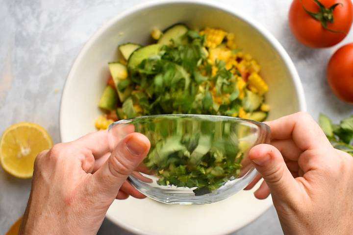 Tomatoes, cucumbers, avocado, and corn being tossed together in a bowl.