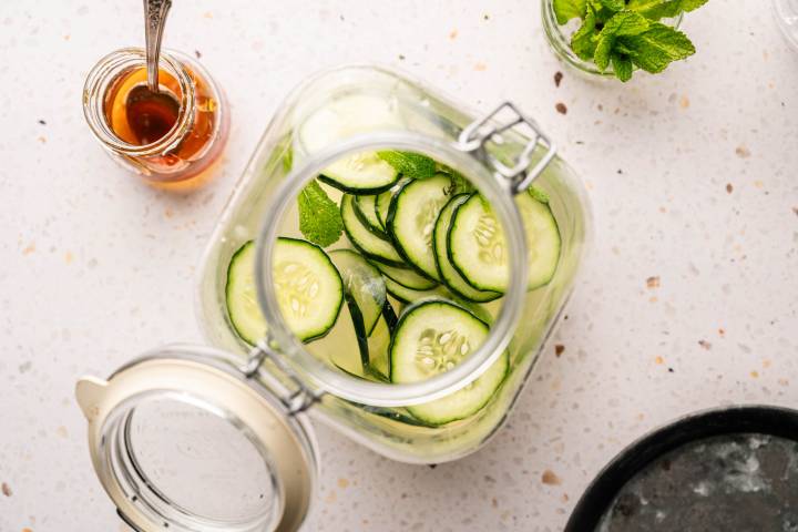 Overhead view of a jar with cucumber slices and mint leaves on a white speckled surface. Nearby are a jar of honey with a spoon and a sprig of mint