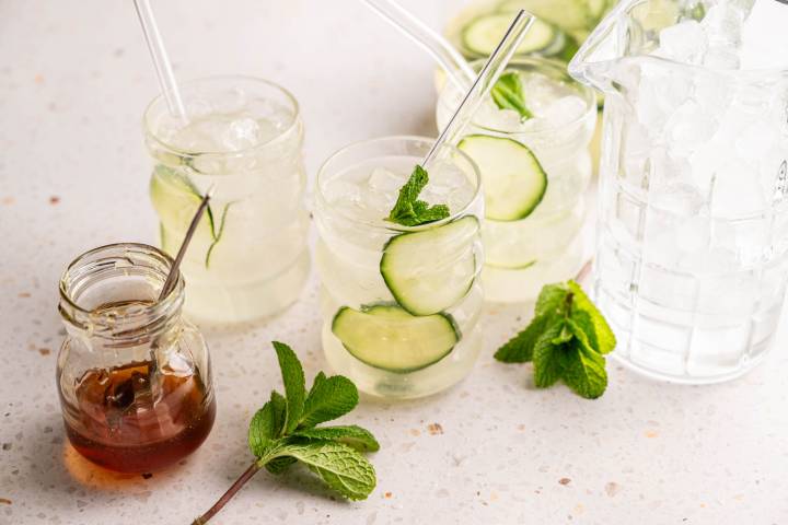 Chilled cucumber mint drinks in glasses with straws, surrounded by a jar of honey, mint leaves, and a pitcher of ice on a speckled countertop. 