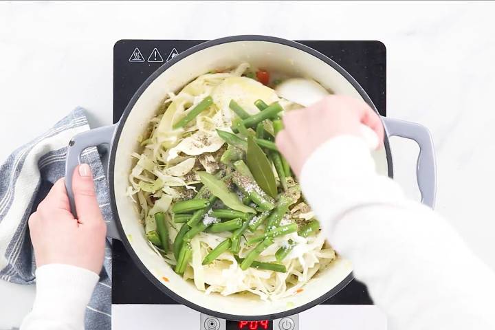 Cabbage, green beans, Italian seasoning, salt, pepper, and bay leaves being stirred to make vegetable soup.