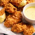 Crispy, golden-brown chicken pieces with mustard coating on a white plate, accompanied by a bowl of creamy lemon-colored dipping sauce.