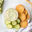 Avocado ranch dip with Greek yogurt served in a small bowl with sliced cucumbers and crackers.