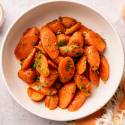 A bowl of glazed carrot slices garnished with parsley, on a white surface. A small bowl with a spoon and a draped cloth are nearby.